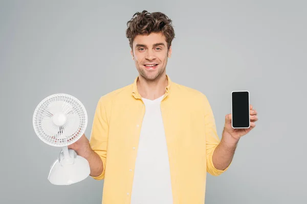 Front view of man smiling, looking at camera and showing smartphone with desk fan isolated on grey — Stock Photo