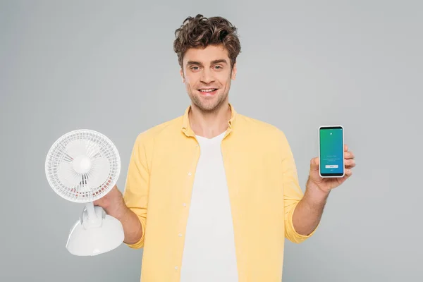 KYIV, UKRAINE - MARCH 27, 2020: front view of man smiling, looking at camera and showing desk fan and smartphone with twitter app isolated on grey — Stock Photo