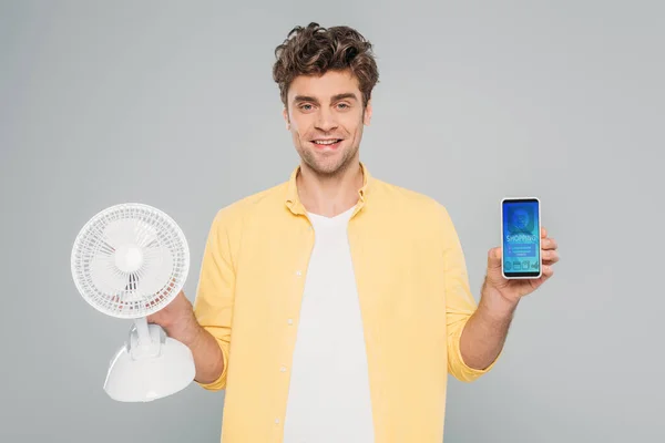 KYIV, UKRAINE - MARCH 27, 2020: front view of man smiling, looking at camera and presenting desk fan and smartphone with shopping app isolated on grey — Stock Photo