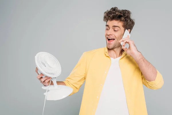 Frontansicht eines aufgeregten Mannes, der auf den Tischventilator blickt und auf dem Smartphone isoliert auf grau spricht — Stockfoto