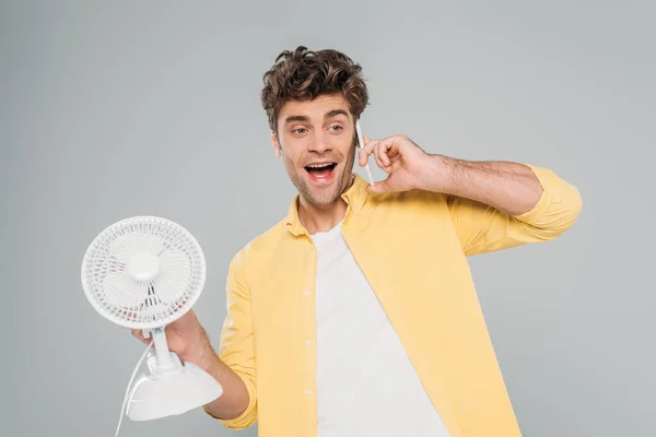 Front view of excited man holding desk fan and talking on smartphone isolated on grey — Stock Photo