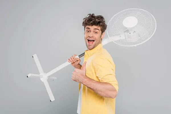 Excited man with open mouth holding electric fan, looking at camera and showing like sign isolated on grey — Stock Photo