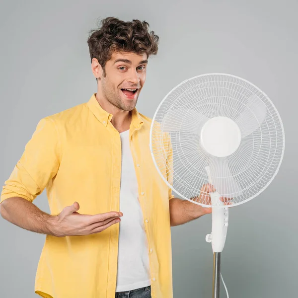 Hombre excitado con la boca abierta mirando a la cámara y apuntando al ventilador eléctrico aislado en gris - foto de stock