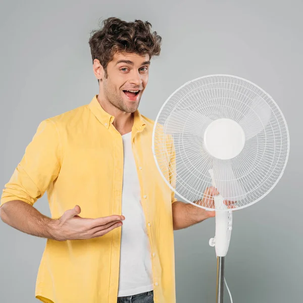 Homem excitado com a boca aberta olhando para a câmera e apontando para ventilador elétrico isolado em cinza — Fotografia de Stock