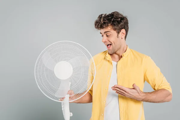 Homem com boca aberta apontando para ventilador elétrico isolado em cinza — Fotografia de Stock