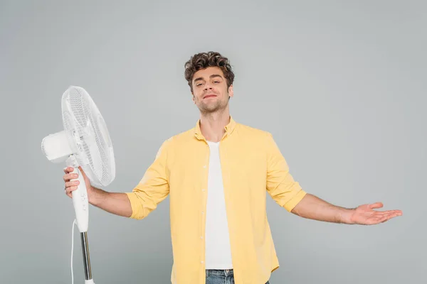 Vorderansicht eines lächelnden Mannes mit offenen Armen in der Nähe eines elektrischen Ventilators isoliert auf grau — Stockfoto