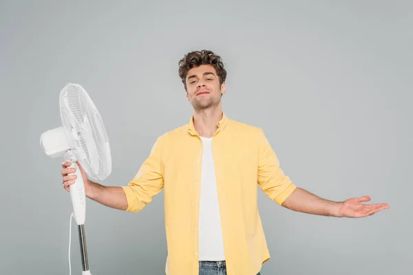 Vista frontale dell'uomo sorridente a braccia aperte vicino ventilatore elettrico isolato su grigio — Foto stock