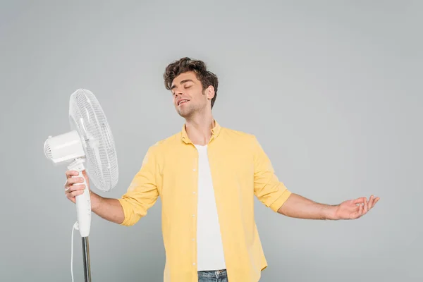 Vorderansicht eines lächelnden Mannes mit geschlossenen Augen und offenen Armen in der Nähe eines elektrischen Ventilators isoliert auf grau — Stockfoto