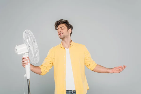 Vista frontal do homem sorrindo com olhos fechados e braços abertos perto de ventilador elétrico isolado em cinza — Fotografia de Stock