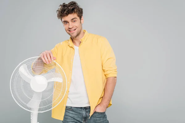 Vista frontal do homem com a mão no bolso sorrindo e olhando para a câmera perto do ventilador elétrico isolado no cinza — Fotografia de Stock