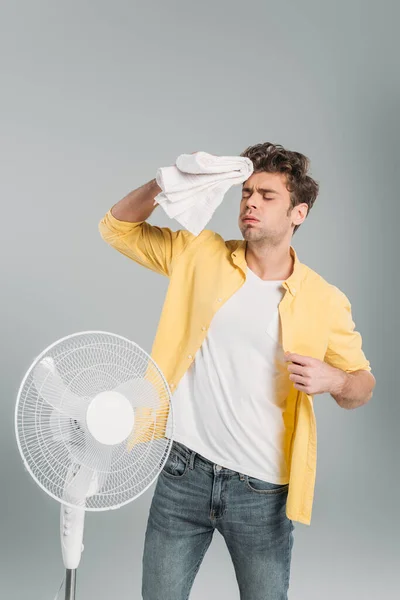 Mann mit Handtuch leidet unter Hitze in der Nähe von Elektroventilator isoliert auf grau — Stockfoto