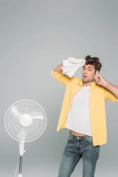 Hombre con toalla mirando a la cámara y hablando en smartphone cerca de ventilador eléctrico aislado en gris - foto de stock