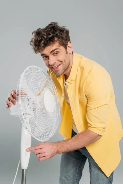 Homem acendendo ventilador elétrico, sorrindo e olhando para a câmera isolada em cinza — Fotografia de Stock