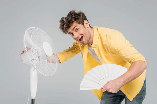 Uomo eccitato con ventilatori elettrici e a mano sorridente isolato sul grigio — Foto stock