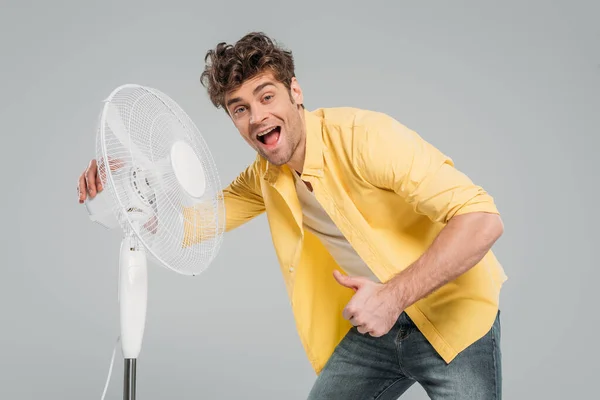 Hombre excitado con la boca abierta cerca de ventilador eléctrico mostrando como signo y mirando a la cámara aislada en gris - foto de stock