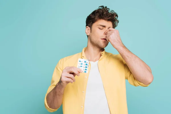 Vista frontale dell'uomo con gli occhi chiusi che toccano il naso e mostrano blister con pillole isolate sul blu — Foto stock