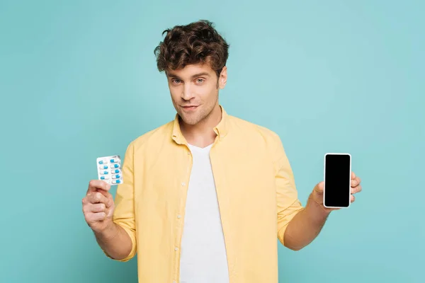 Vista frontal del hombre sonriendo, mirando a la cámara y mostrando el teléfono inteligente y el paquete de ampollas con pastillas aisladas en azul - foto de stock