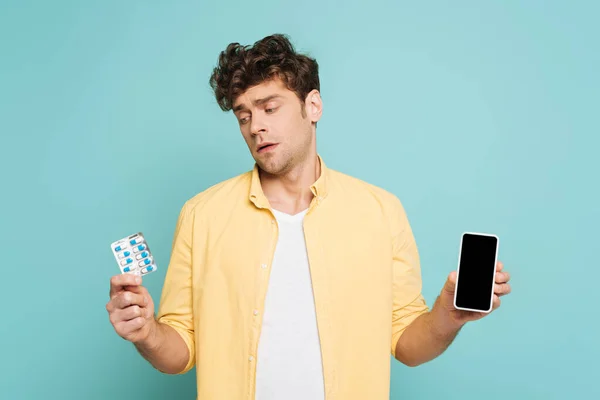 Frontansicht eines Mannes, der Blisterverpackung mit Pillen betrachtet und Smartphone isoliert auf blauem Grund zeigt — Stockfoto