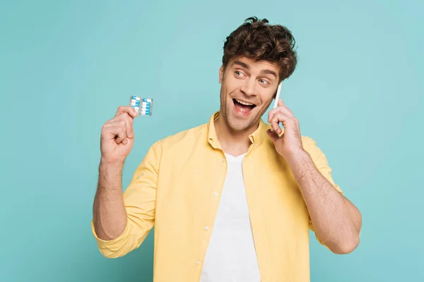 Вид спереди возбужденного человека, говорящего по смартфону и держащего пузырьковый пакет с таблетками, изолированными синим цветом — стоковое фото