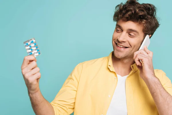 Hombre sonriendo, hablando por teléfono inteligente y sosteniendo blíster con pastillas aisladas en azul - foto de stock