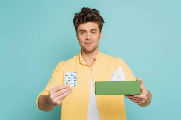 Вид спереди мужчины, показывающего пузырьковый пакет с таблетками и коробку с салфетками и смотрящего на камеру, изолированную на синем фоне — стоковое фото