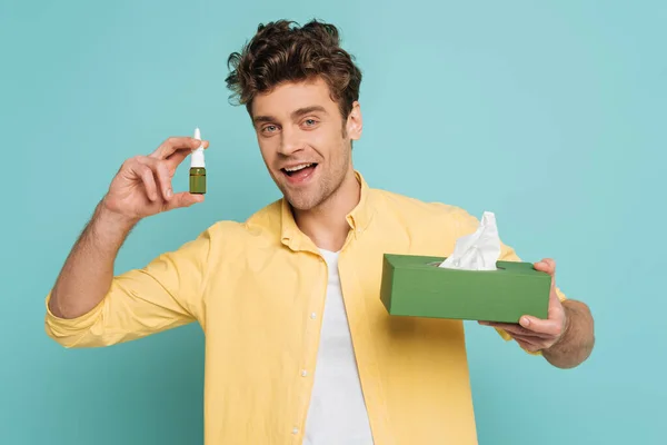 Vista frontal del hombre sonriendo y mostrando botella con gotas nasales y caja con servilletas aisladas en azul - foto de stock