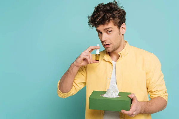 Mann hält Schachtel mit Servietten, blickt in Kamera und benutzt Nasentropfen isoliert auf blauem Grund — Stockfoto