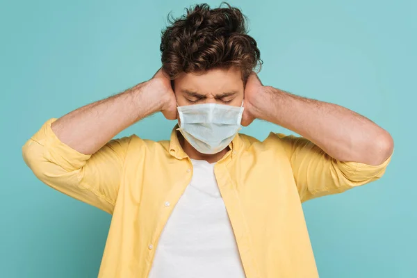 Vista frontal del hombre en máscara médica con los ojos cerrados cubriendo las orejas aisladas en azul - foto de stock