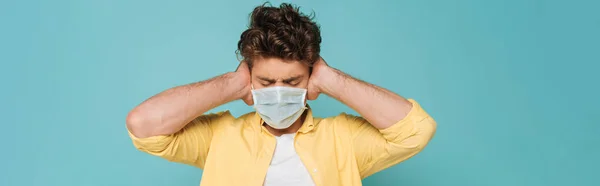 Vista frontal del hombre en máscara médica con los ojos cerrados cubriendo las orejas aisladas en azul, tiro panorámico - foto de stock