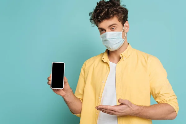 Человек в медицинской маске указывает на смартфон, изолированный на голубом — стоковое фото