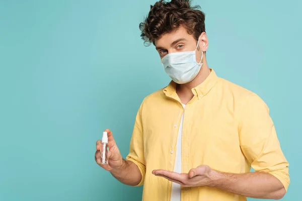 Uomo in maschera medica che punta alla bottiglia di disinfettante per le mani isolato su blu — Foto stock