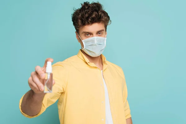 Homme en masque médical regardant la caméra et montrant bouteille de désinfectant pour les mains isolé sur bleu — Photo de stock