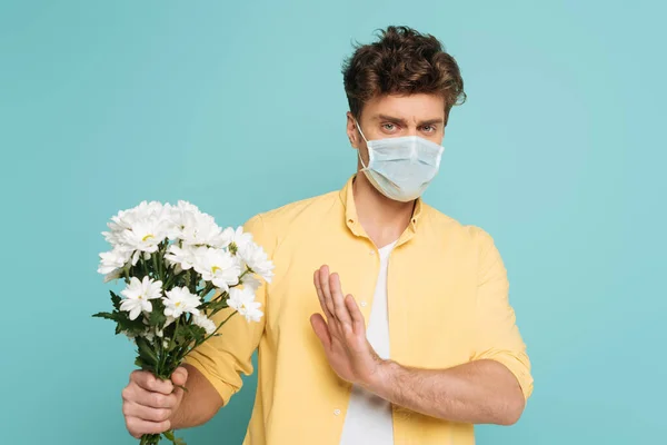 Hombre en máscara médica con la mano extendida sosteniendo ramo y mostrando ningún signo aislado en azul - foto de stock