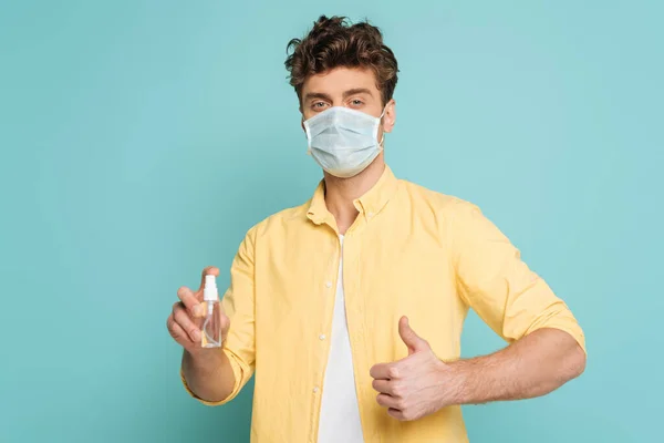 Mann in medizinischer Maske hält Flasche mit Händedesinfektionsmittel und zeigt wie ein Zeichen isoliert auf blauem Grund — Stockfoto