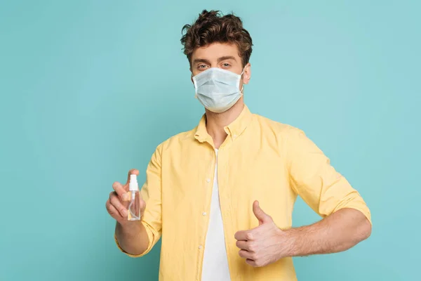 Hombre con máscara médica sosteniendo botella de desinfectante de manos y mostrando como signo aislado en azul - foto de stock