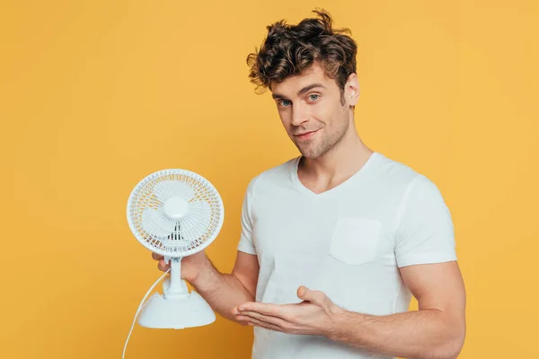 Mann lächelt, blickt in Kamera und zeigt auf gelb isolierten Tischventilator — Stockfoto