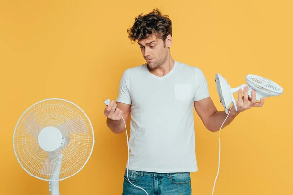 Vista frontal do homem com ventiladores elétricos e de mesa olhando para ficha elétrica isolada em amarelo — Fotografia de Stock