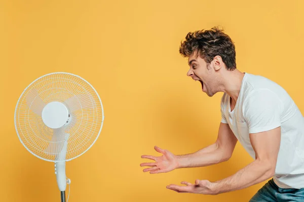 Excited man shouting with outstretched hands in front of electric fan on yellow — Stock Photo