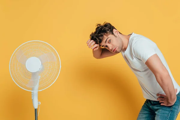 Homem com a mão no quadril olhando para a câmera e inclinando-se perto do ventilador elétrico no amarelo — Fotografia de Stock