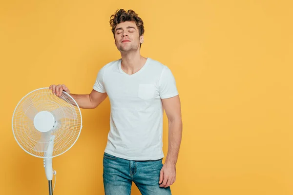 Vista frontal do homem alegre com os olhos fechados tocando ventilador elétrico no amarelo — Fotografia de Stock