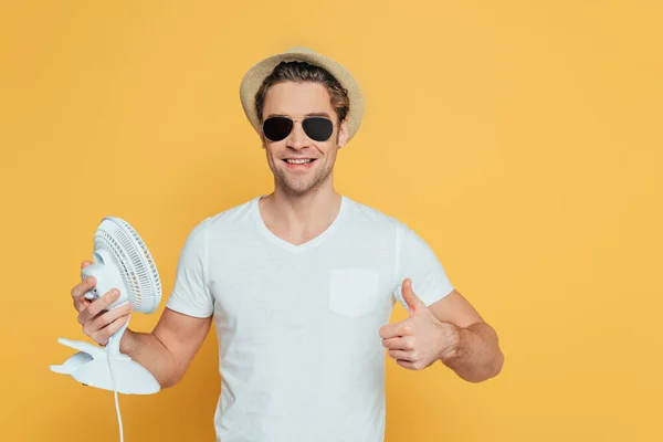 Вид спереди человека в шляпе и солнцезащитных очках, держащего настольный вентилятор и показывающего знак, изолированный на желтом — стоковое фото