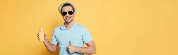 Frontansicht eines Mannes mit Hut und Sonnenbrille mit einer Flasche Sonnencreme, die lächelt und wie ein Schild auf gelbem, panoramischen Foto zeigt — Stockfoto