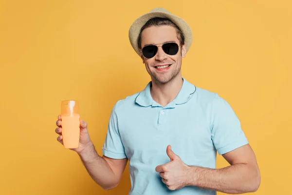 Front view of man in hat and sunglasses with bottle of sunscreen smiling and showing like sign isolated on yellow — Stock Photo