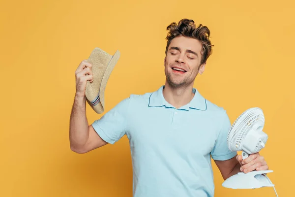 Vue de face de l'homme joyeux avec les yeux fermés souriant et tenant chapeau avec ventilateur de bureau isolé sur jaune — Photo de stock