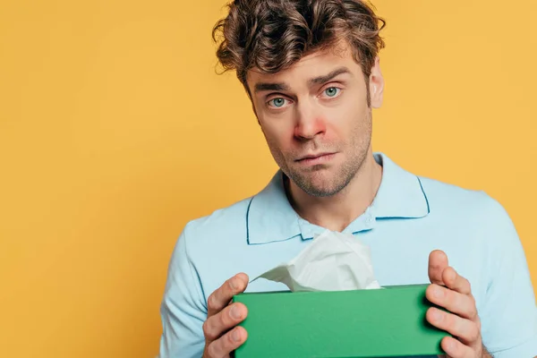 Triste y enfermo hombre mostrando caja con servilletas y mirando a la cámara aislada en amarillo - foto de stock