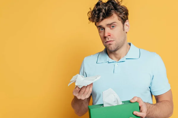 Triste y enfermo hombre mostrando caja con servilletas y mirando a la cámara aislada en amarillo - foto de stock