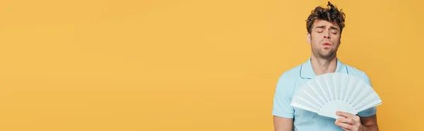 Front view of man with closed eyes and hand fan suffering from heat isolated on yellow, panoramic shot — Stock Photo