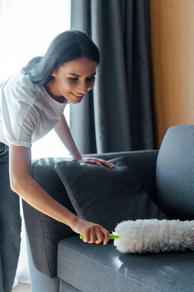 Glückliche schöne Frau putzt Staub vom Sofa mit Staubwedel — Stockfoto