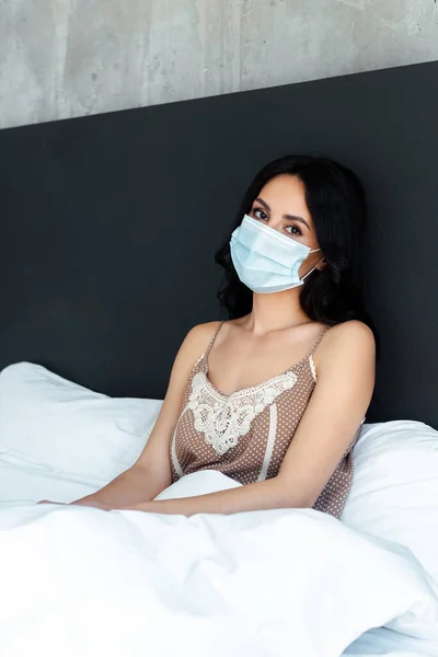 Привлекательная женщина в медицинской маске сидит в постели дома на самоизоляции — стоковое фото