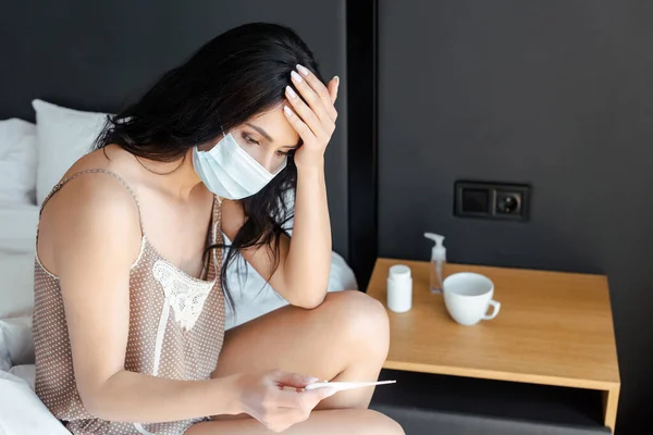 Preocupada chica enferma en máscara médica mirando el termómetro mientras está sentada en la cama en el auto aislamiento - foto de stock